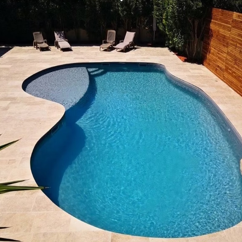 Piscine de forme libre bleue avec plage immergée, terrasse en travertin clair et chaises longues de jardin.