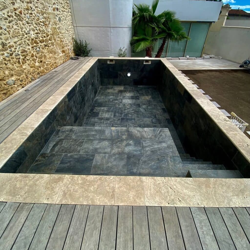 Piscine moderne vide avec revêtement en ardoise sombre, margelles en travertin et terrasse en bois.