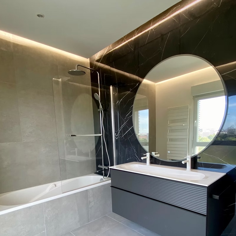 Salle de bain design : baignoire, double vasque et grand miroir rond rétroéclairé sur mur en marbre noir.