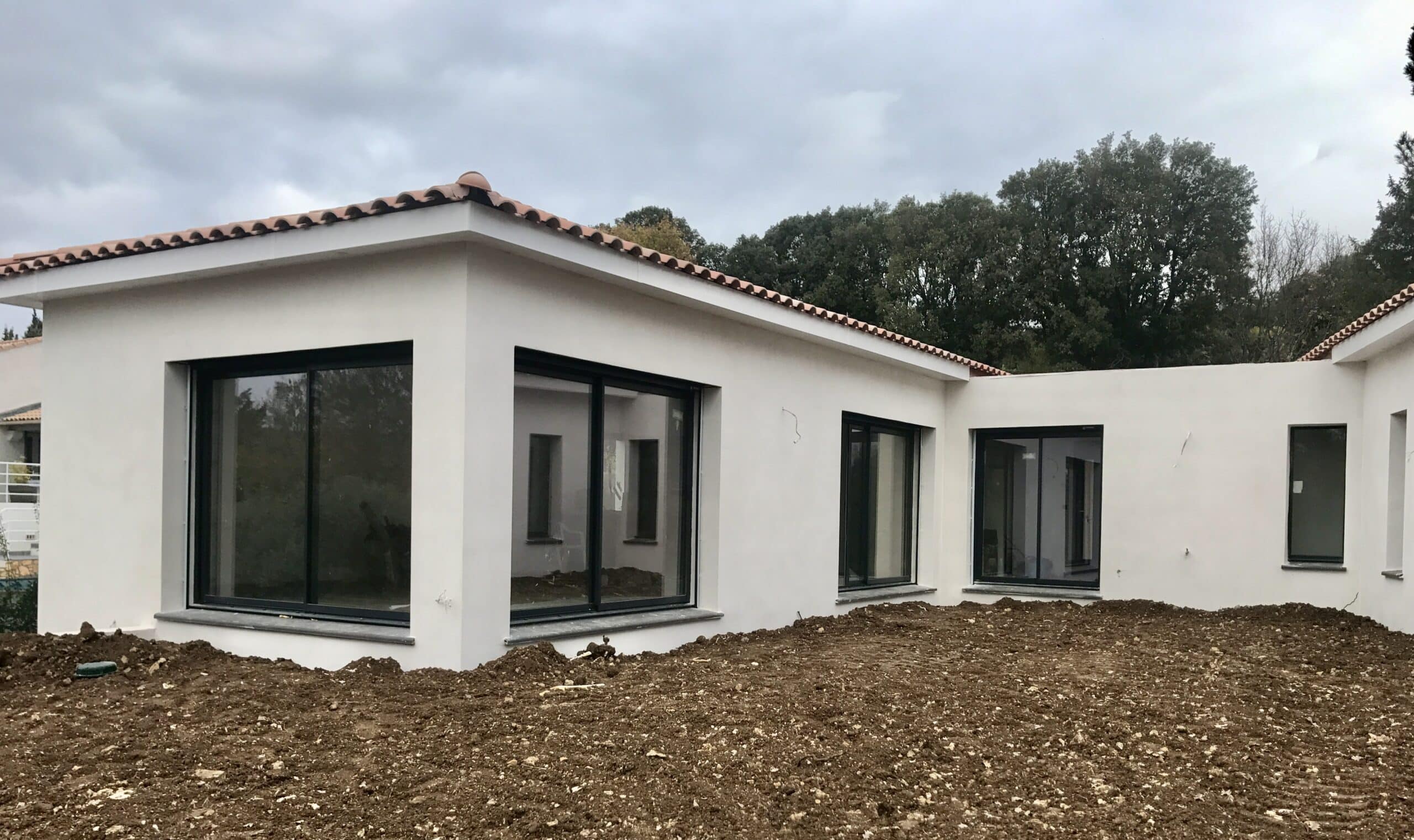 Maison Moderne : Façade Blanche et Menuiseries Noires Maison moderne blanche avec menuiseries noires et baies vitrées sur un terrain en cours de construction.