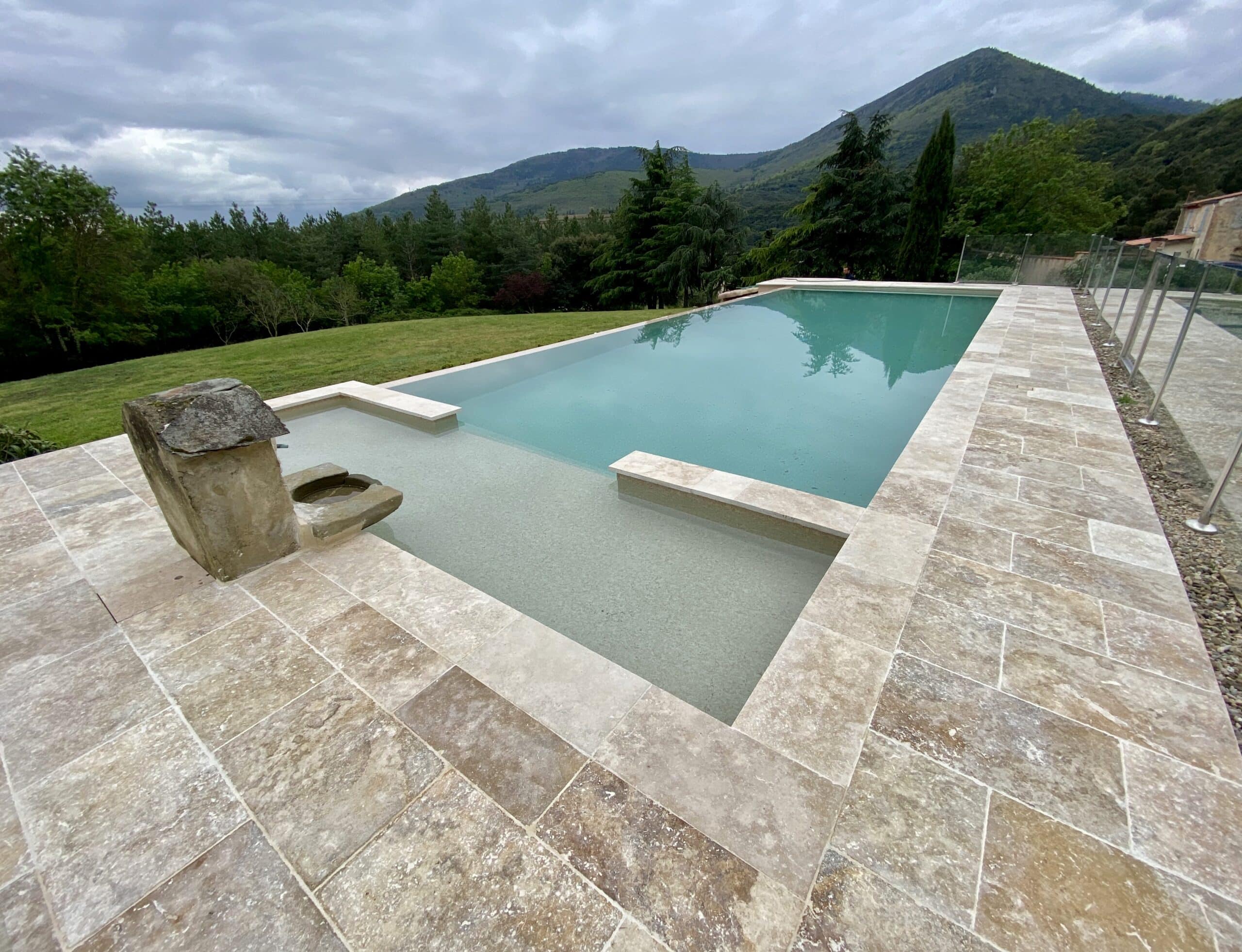 Piscine à débordement moderne et terrasse en travertin Piscine à débordement moderne avec terrasse en travertin devant un paysage de montagnes sous un ciel couvert.