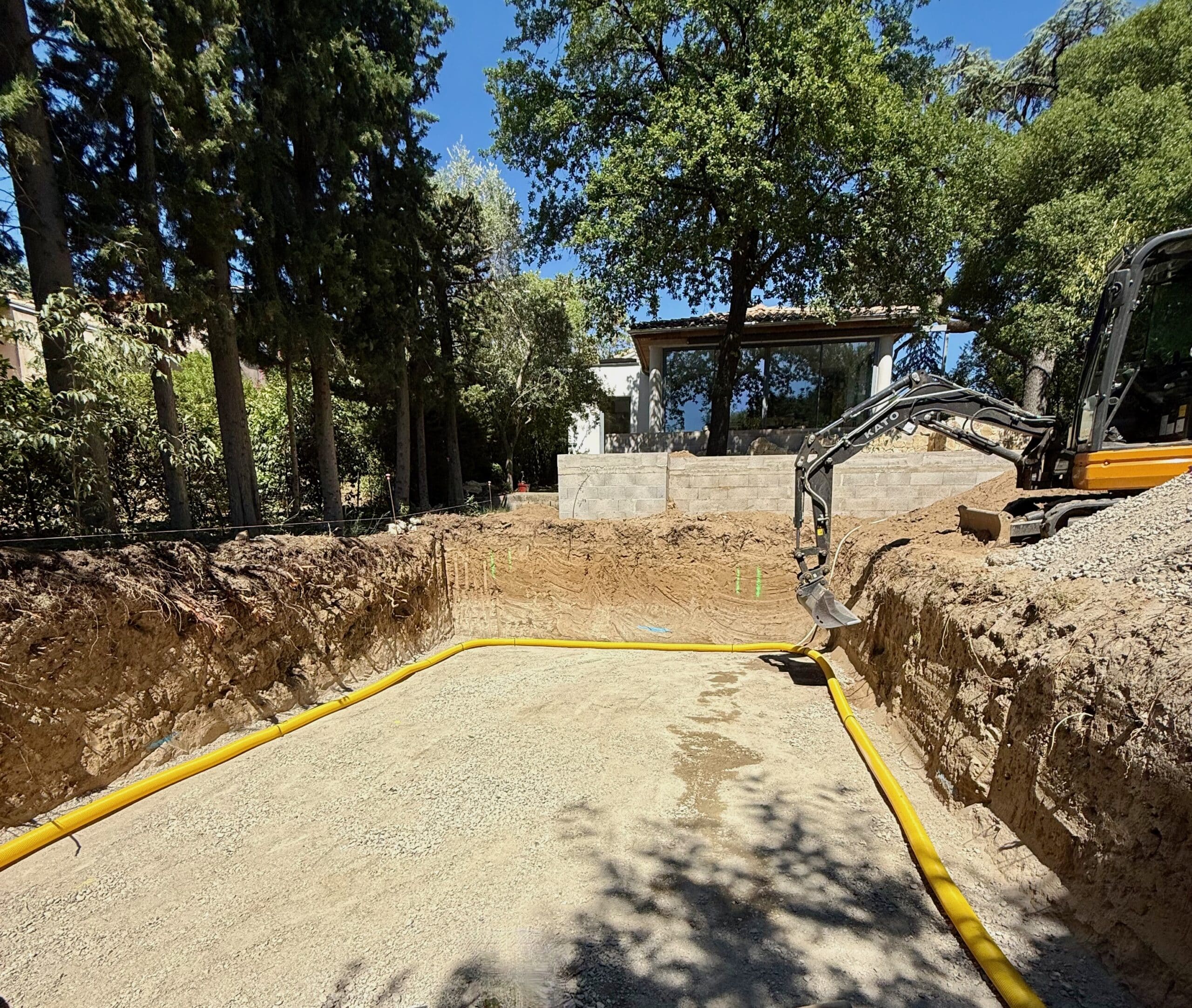 Construction de piscine : étape d’excavation et terrassement Chantier de piscine avec excavation, pelleteuse KATO et tuyau de drainage jaune dans un jardin arboré.