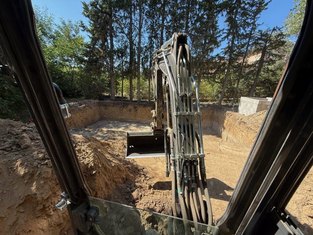 Vue de l'intérieur d'une pelleteuse excavant un terrain pour des fondations devant une rangée d'arbres.