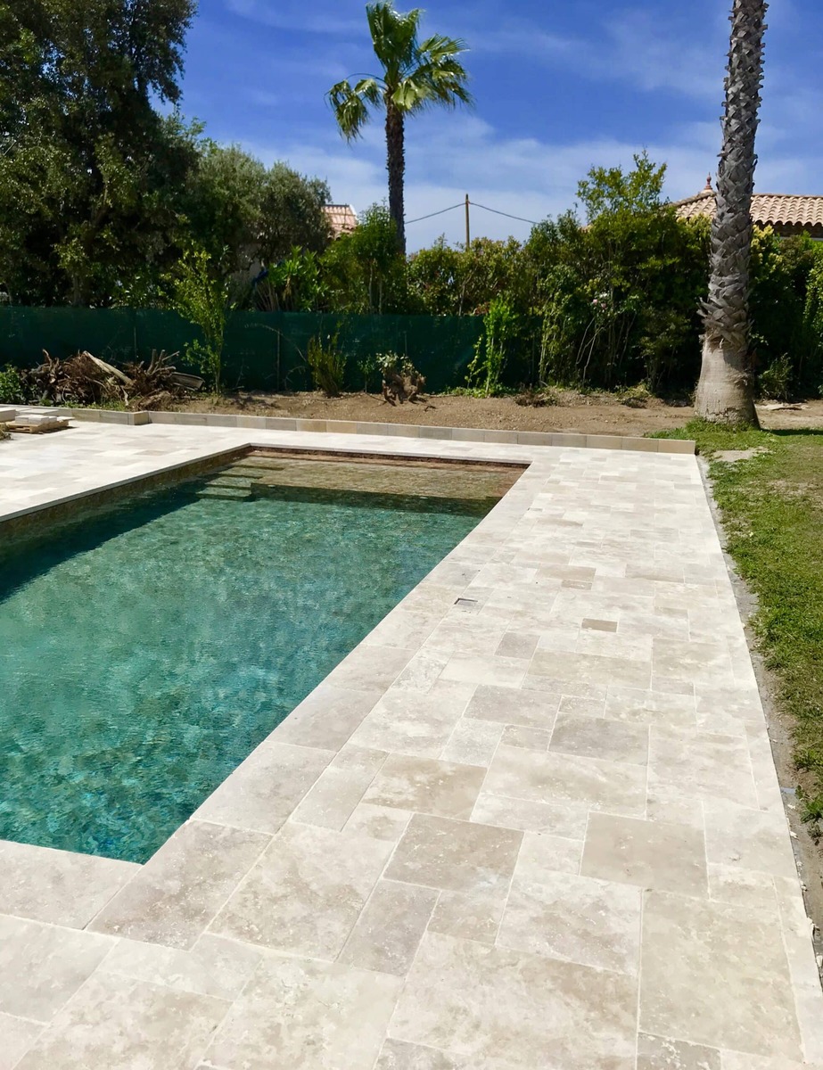 Piscine moderne avec margelles en pierre beige, eau turquoise, jardin ensoleillé et palmiers sous ciel bleu.