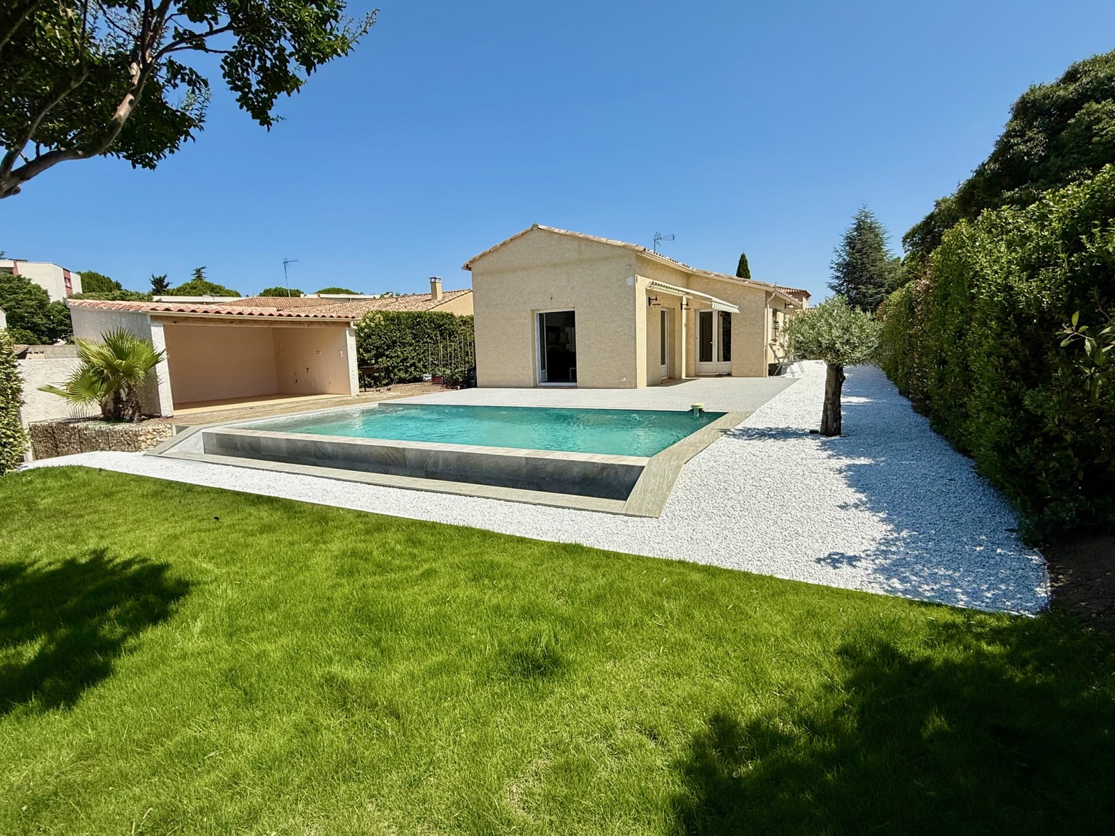 Maison avec piscine, pelouse verte et gravier blanc sous ciel bleu.
