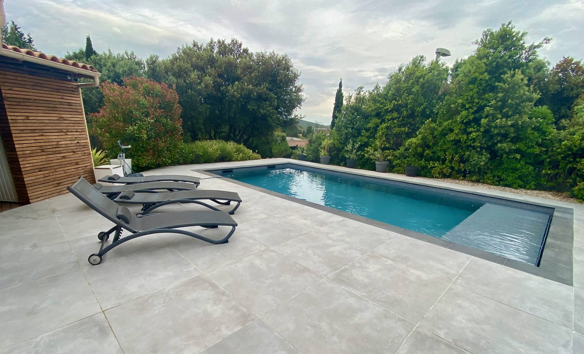 Piscine rectangulaire moderne, terrasse en pierre grise avec transats et abri bois entourés de végétation luxuriante.