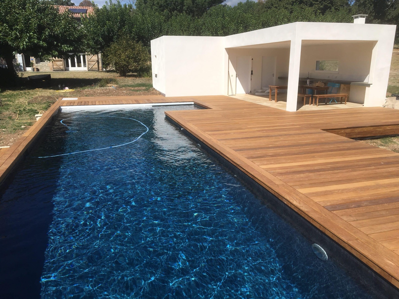 Piscine liner bleu foncé, terrasse en bois et pool house blanc moderne dans un jardin.