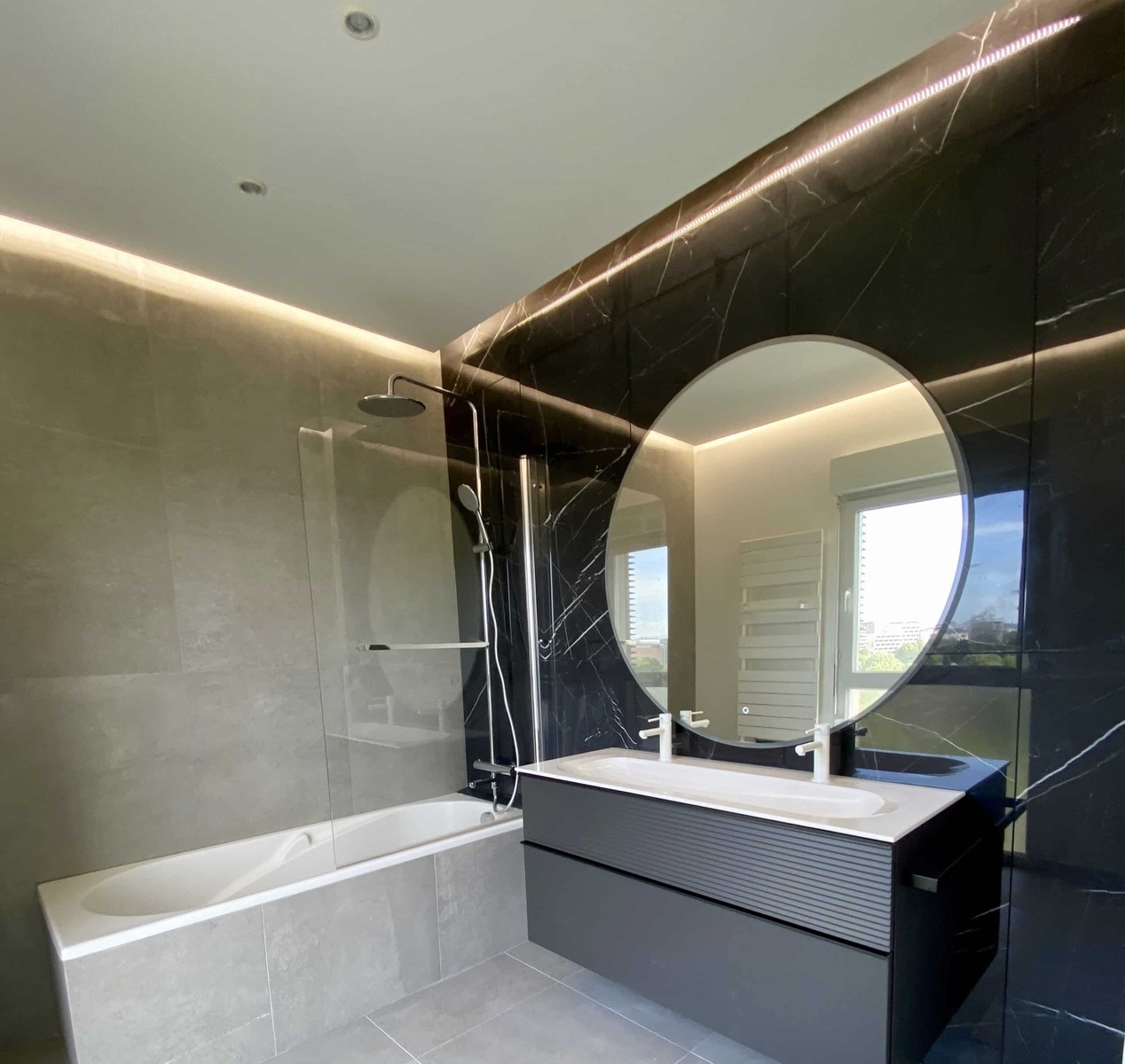 Salle de bain moderne avec baignoire, meuble gris et grand miroir rond sur mur effet marbre noir.