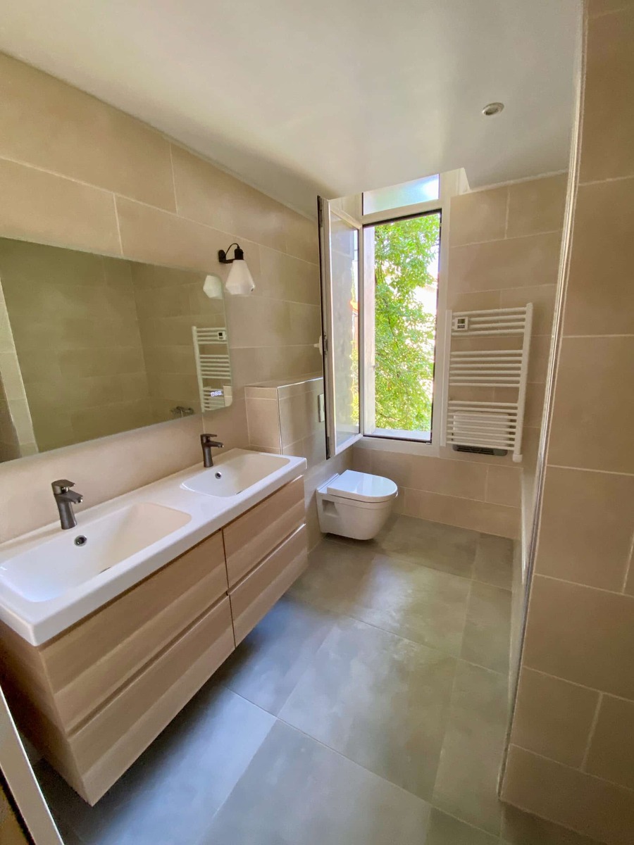 Salle de bain moderne : double vasque bois, WC suspendu et sèche-serviettes blanc sous une fenêtre lumineuse.