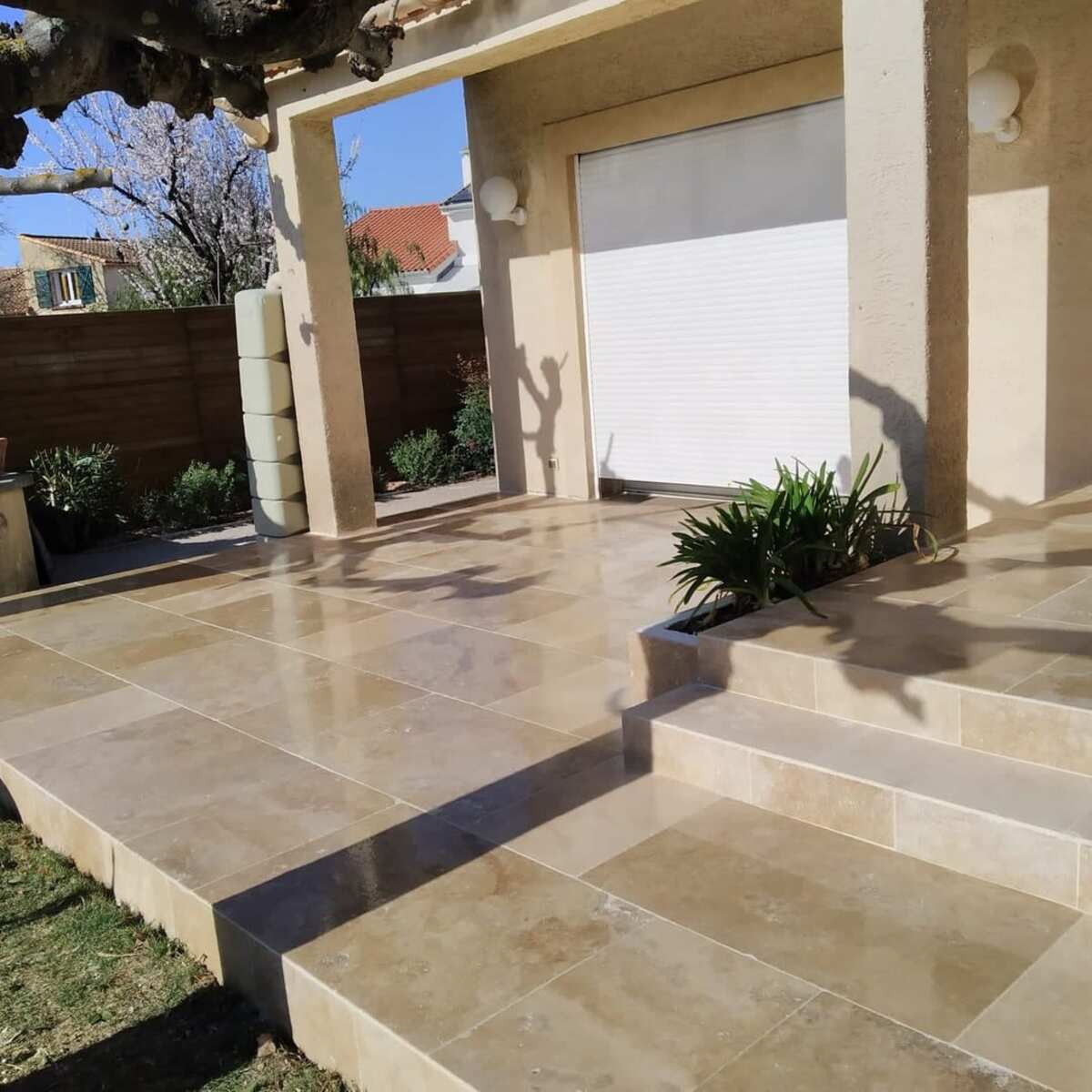 Terrasse en travertin beige brillant avec escalier sous un auvent ensoleillé et jardin.